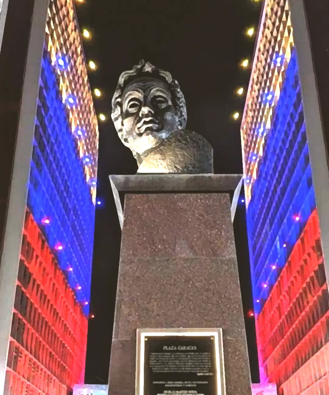 Plaza Caracas at night: a bronze bust on its granite pedestal, between the two lit towers of Centro Simón Bolívar